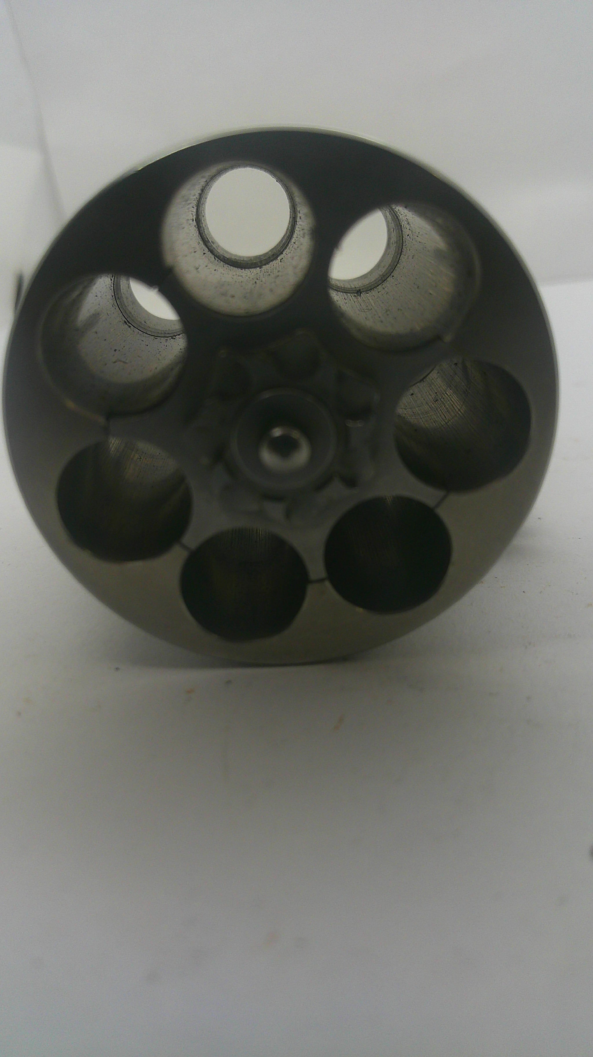 A dark gray metal revolver cylinder or magazine component with six circular chambers, photographed on a plain white background.