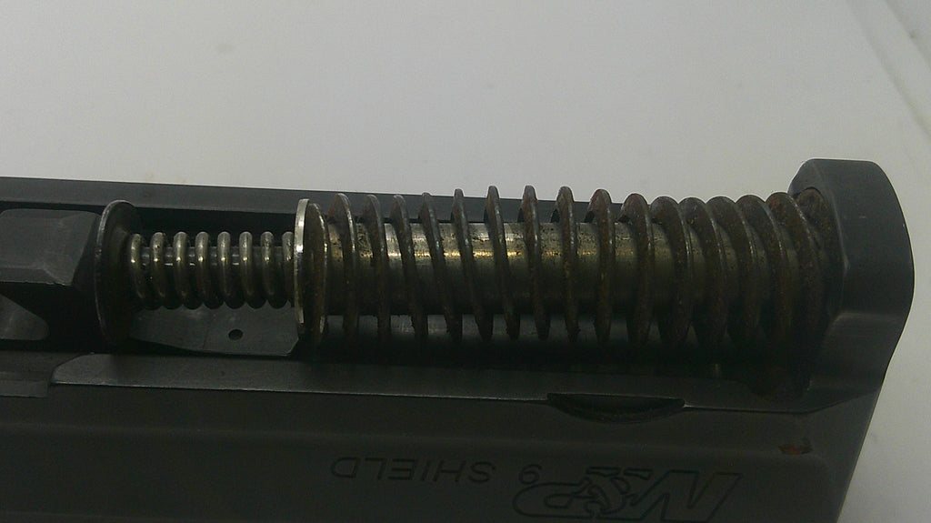 Close-up of a black metal firearm component with a prominent coiled spring and visible 'N&P SHIELD' branding on the side, set against a plain light background.