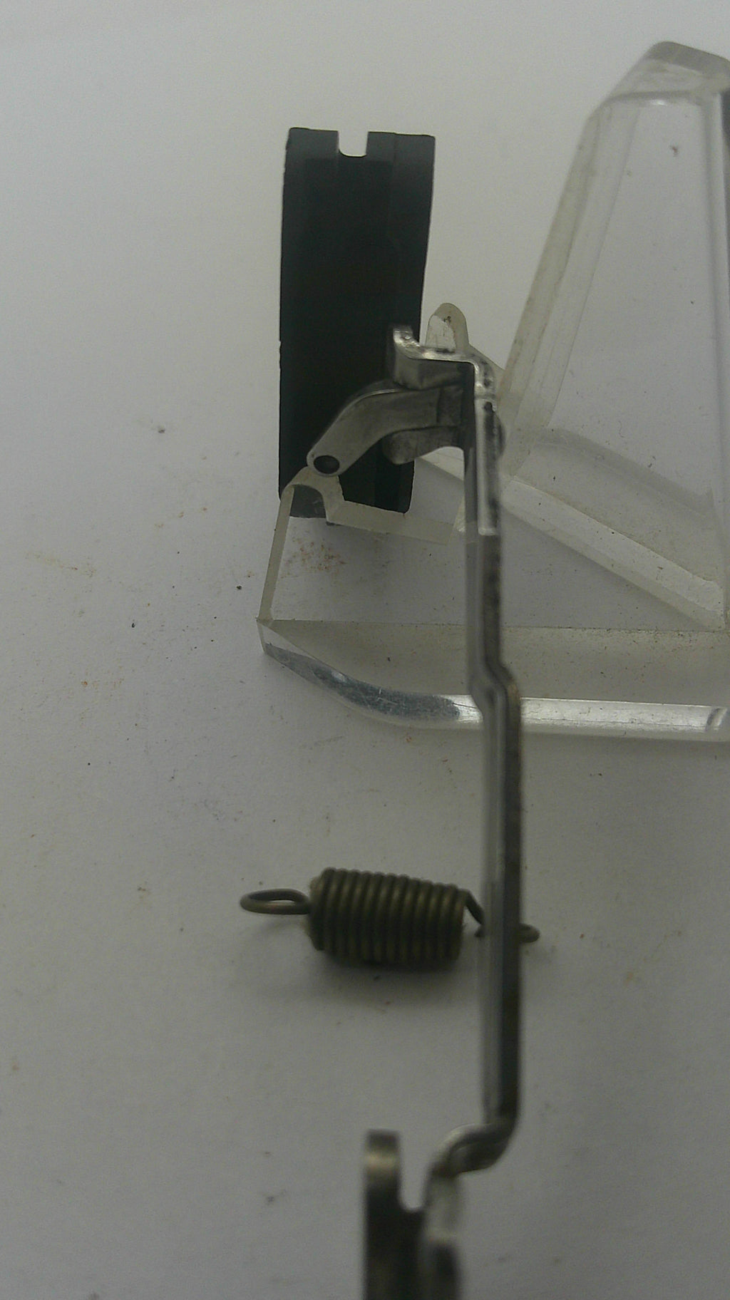 A close-up of a sewing machine needle clamp and spring assembly, showing a metal needle bar, black needle plate, and a small coiled metal spring on a light gray background.