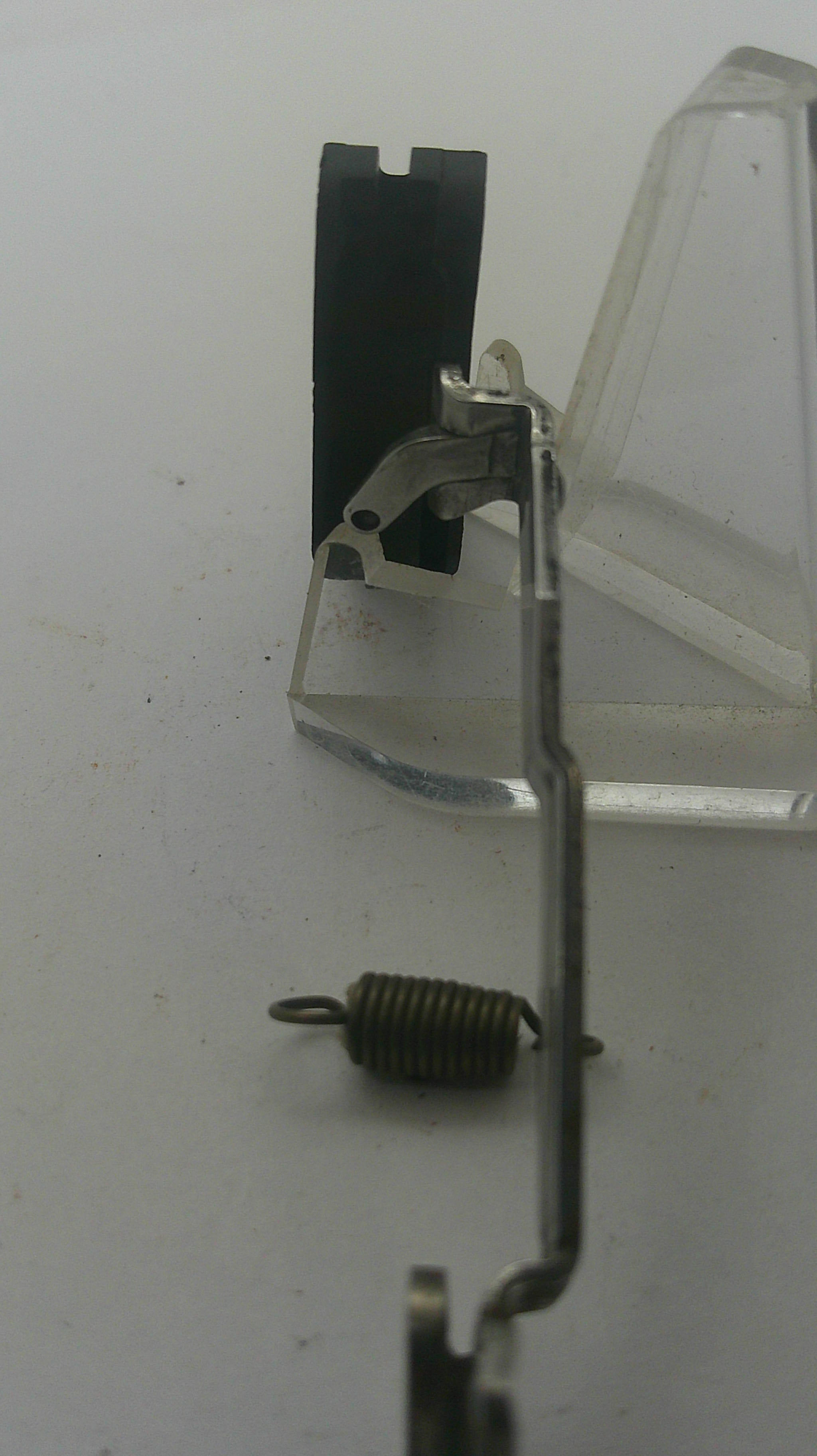 A close-up of a sewing machine needle clamp and spring assembly, showing a metal needle bar, black needle plate, and a small coiled metal spring on a light gray background.