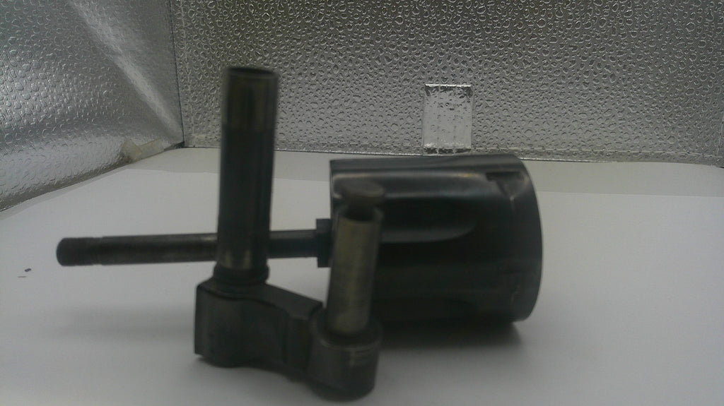 A used black plastic mechanical part, likely a pump or filter assembly, photographed on a white surface with a textured silver background.