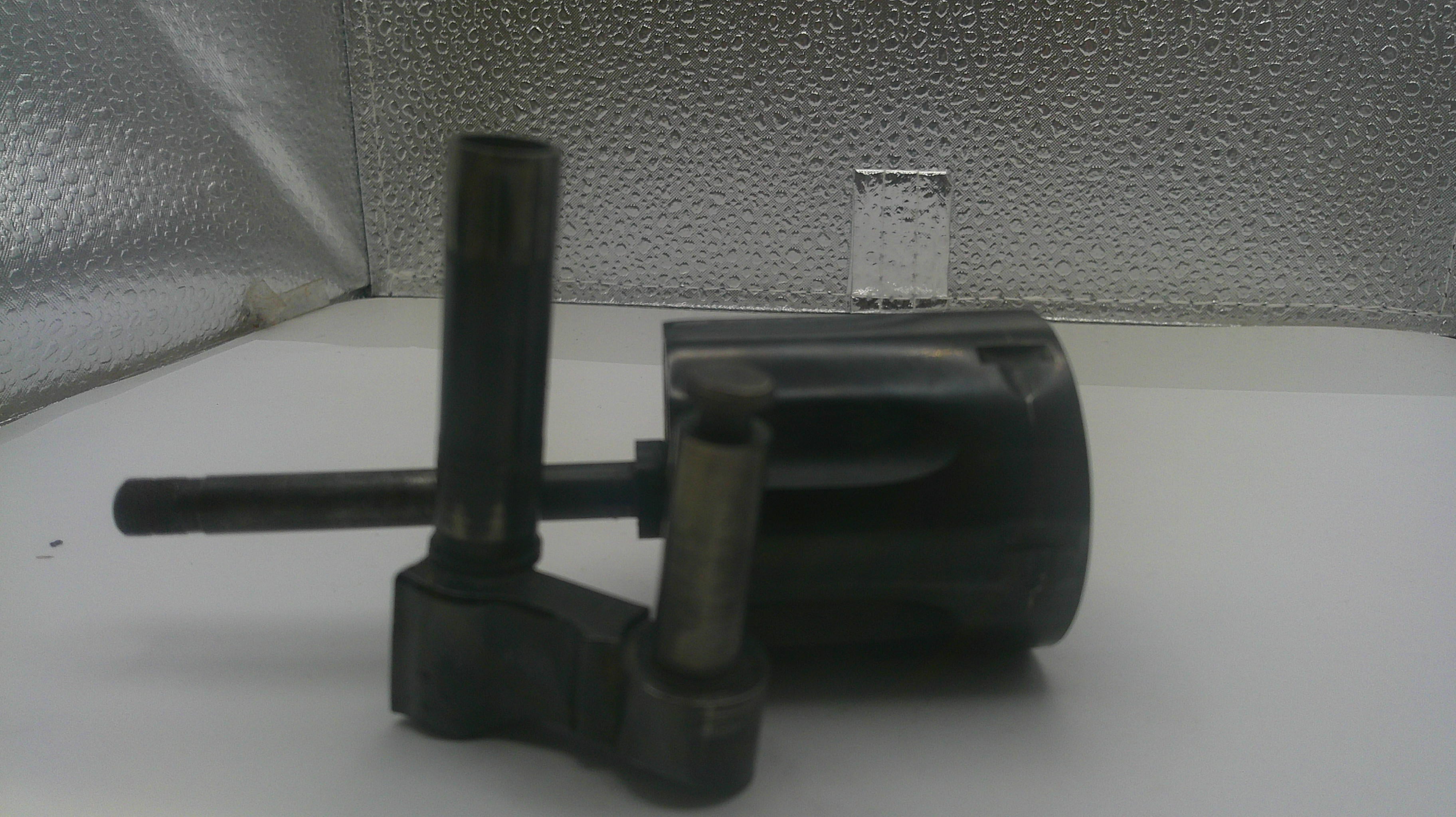 A used black plastic mechanical part, likely a pump or filter assembly, photographed on a white surface with a textured silver background.