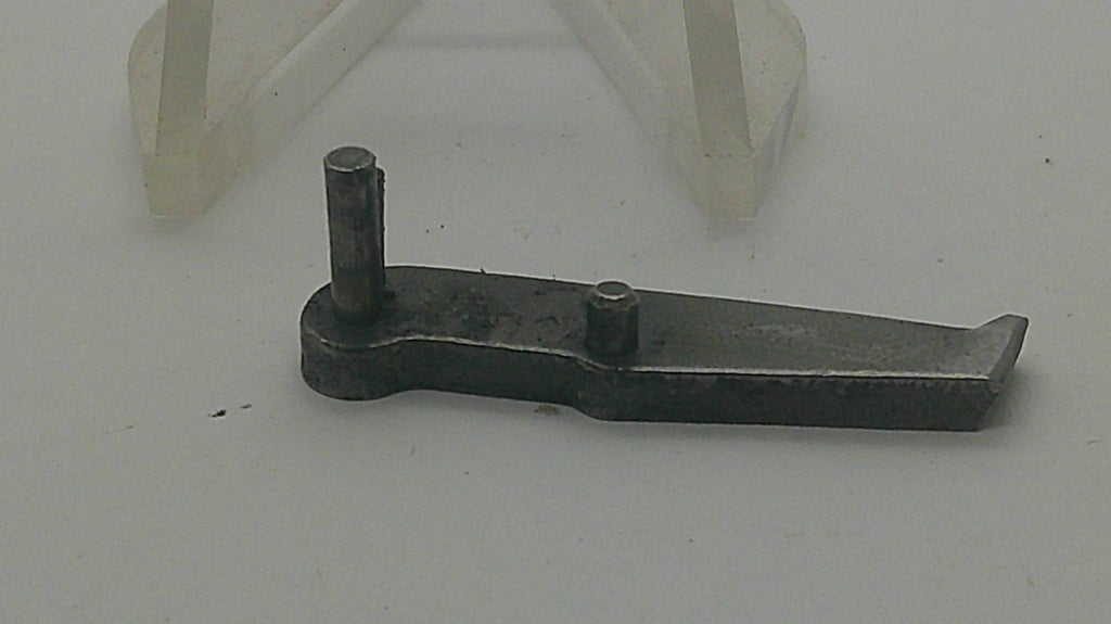 A small dark metal mechanical part with two vertical pins, photographed on a light gray background with a translucent plastic stand visible behind it.