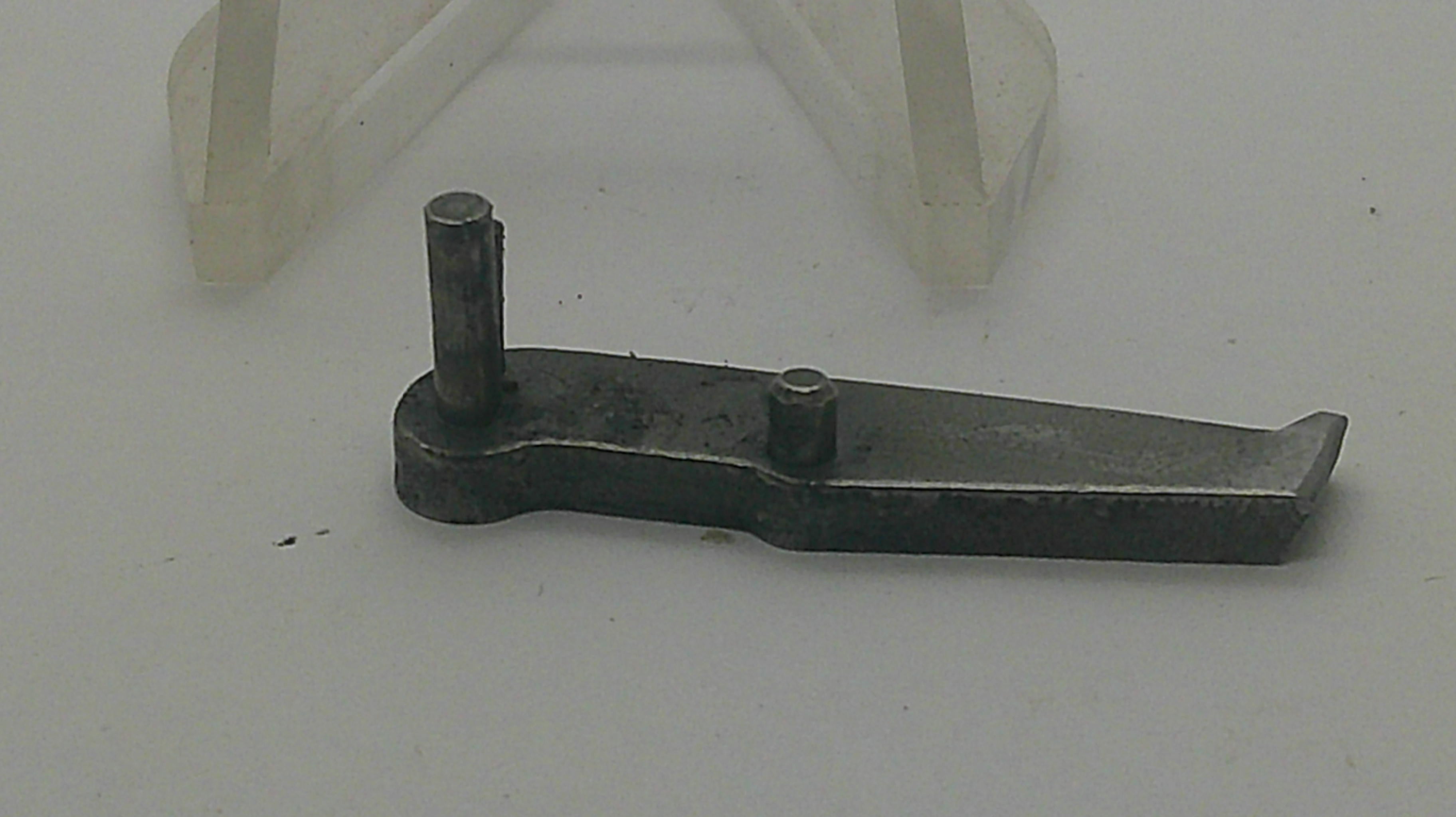 A small dark metal mechanical part with two vertical pins, photographed on a light gray background with a translucent plastic stand visible behind it.