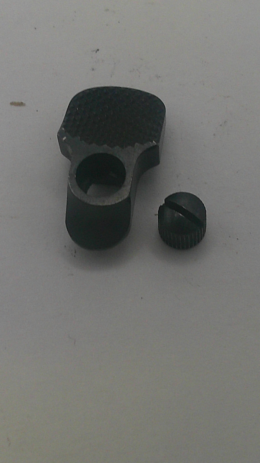 A small black metal hardware component with a textured knurled side and a separate matching cap or knob, placed on a plain light gray surface.