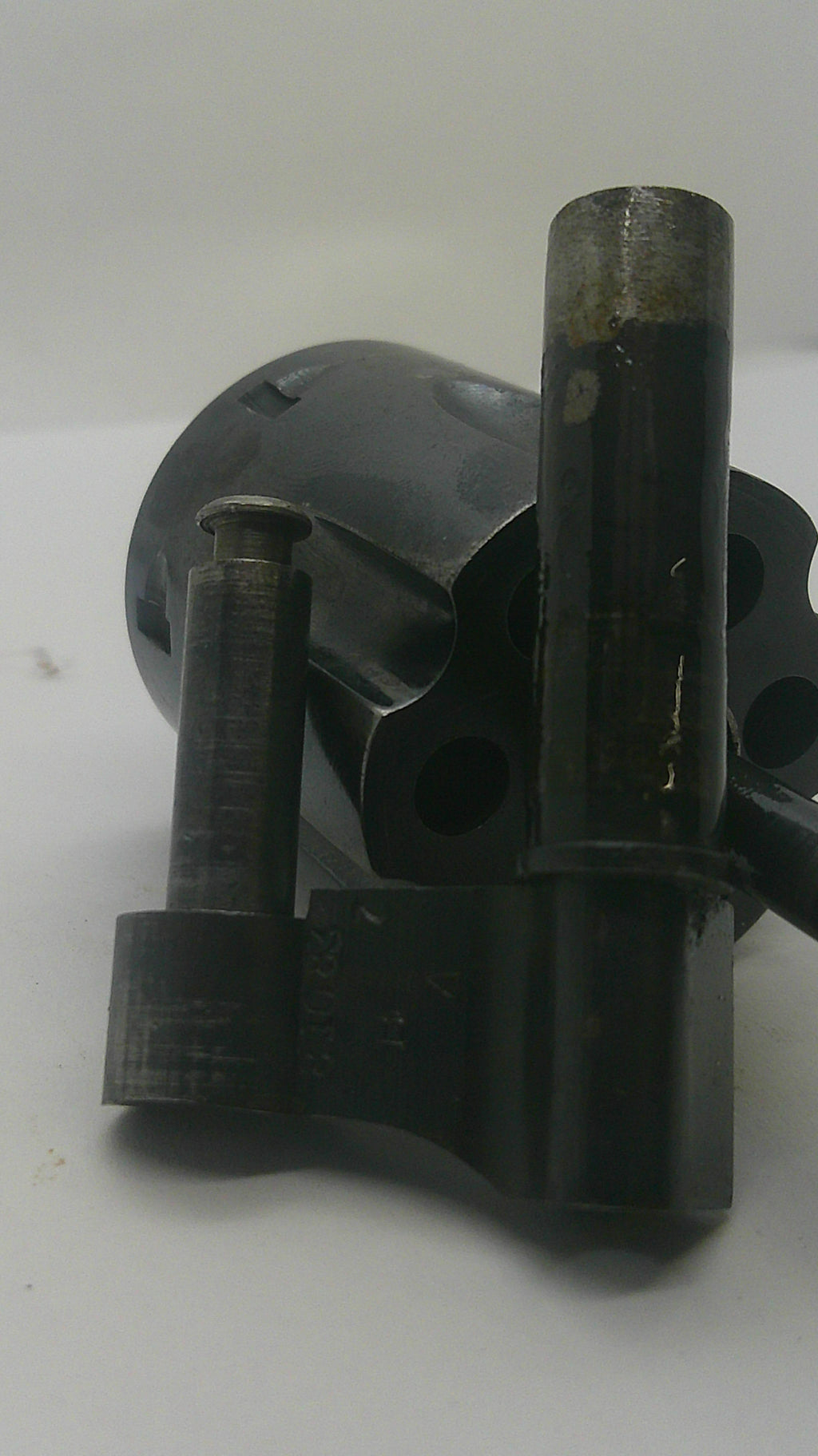 A close-up of a dark metal mechanical part or tool component with cylindrical rods and circular openings, photographed against a plain light background.