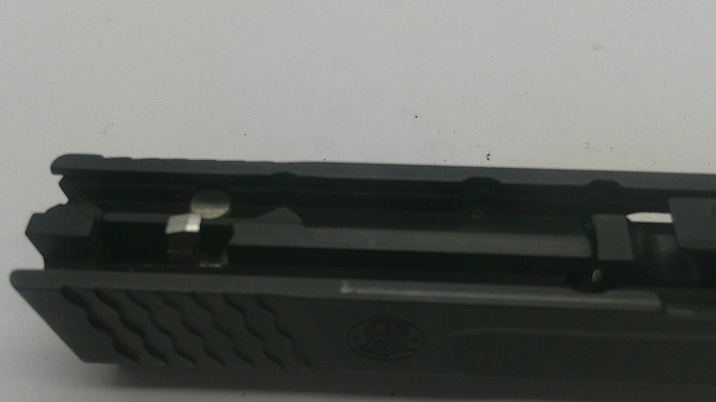 A close-up of a black textured firearm grip or handle component with a metallic lever and a small embossed logo, photographed against a plain light background.