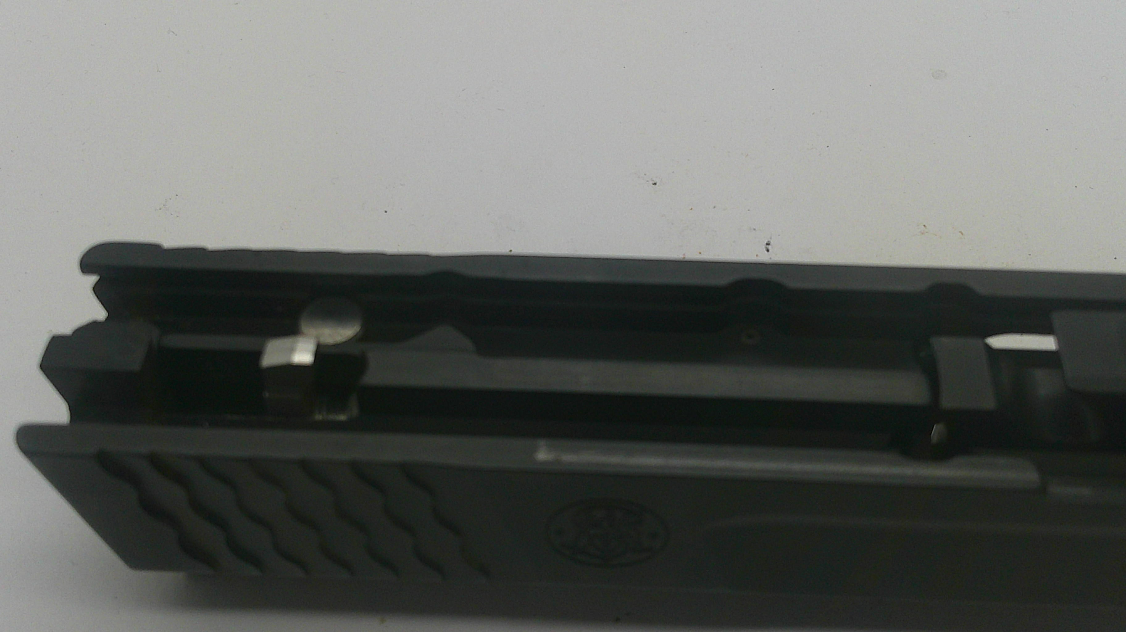 A close-up of a black textured firearm grip or handle component with a metallic lever and a small embossed logo, photographed against a plain light background.