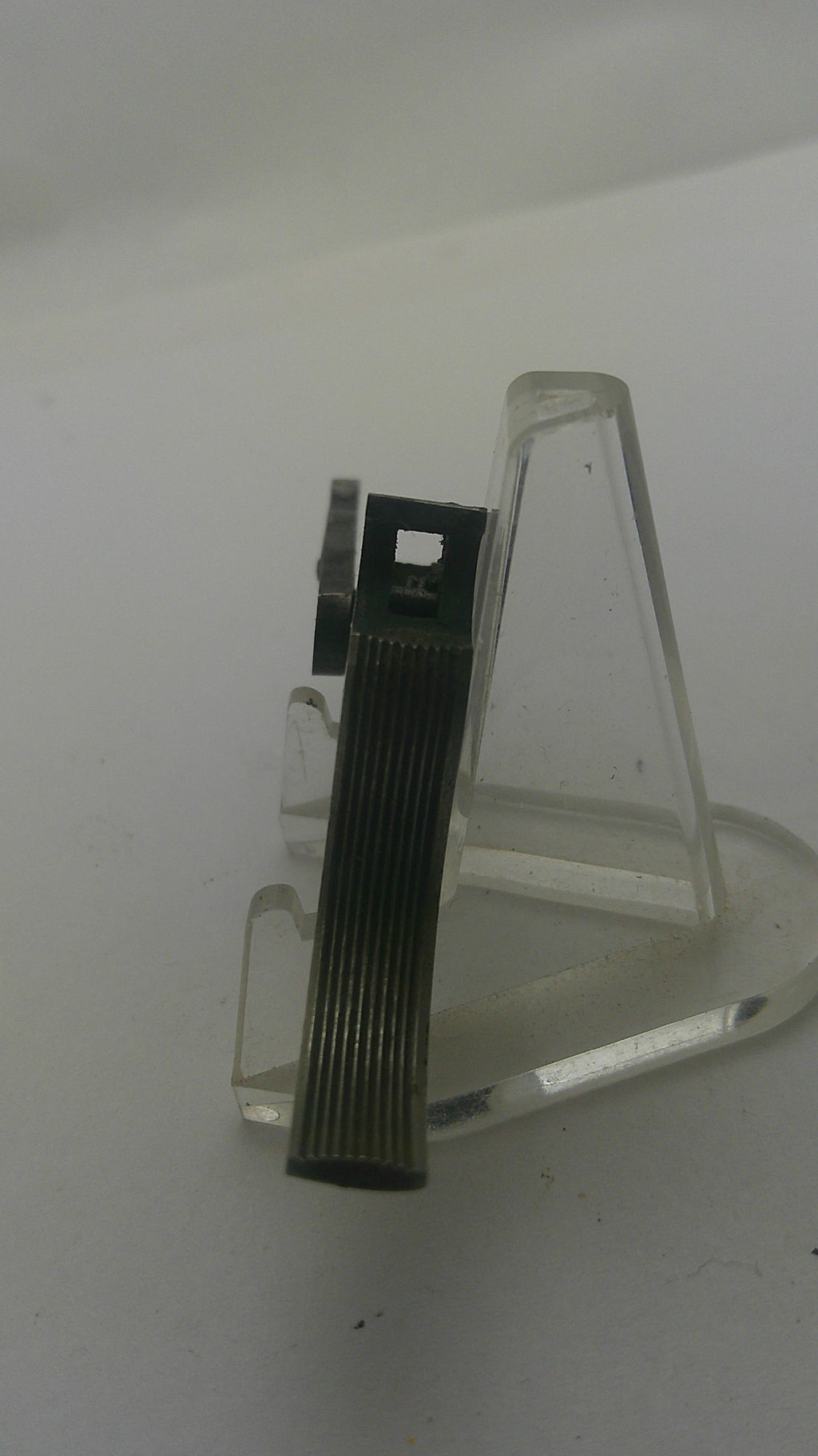 A small black ribbed rectangular clip or fastener with a small metal bracket, displayed on a clear plastic stand against a plain light background.