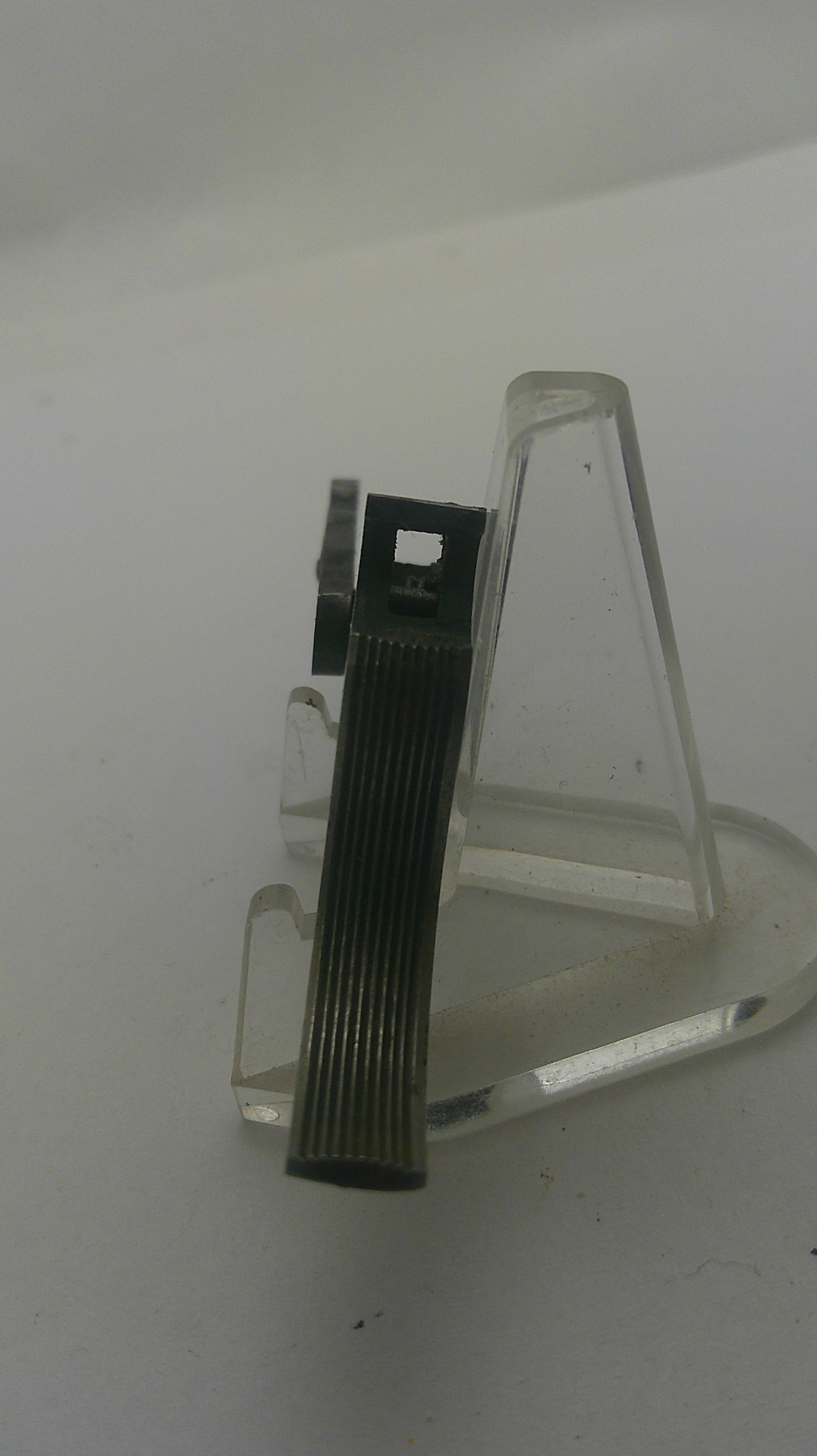 A small black ribbed rectangular clip or fastener with a small metal bracket, displayed on a clear plastic stand against a plain light background.
