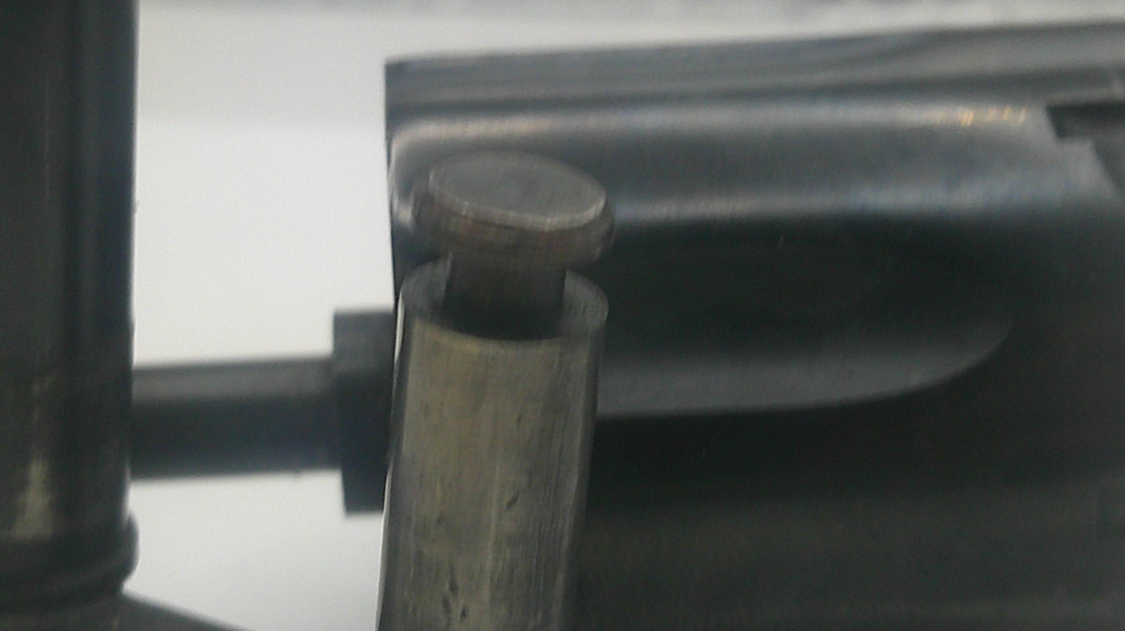 Close-up of a dark metal cylindrical component with a round metallic cap or knob, set against a mostly dark and light gray background.