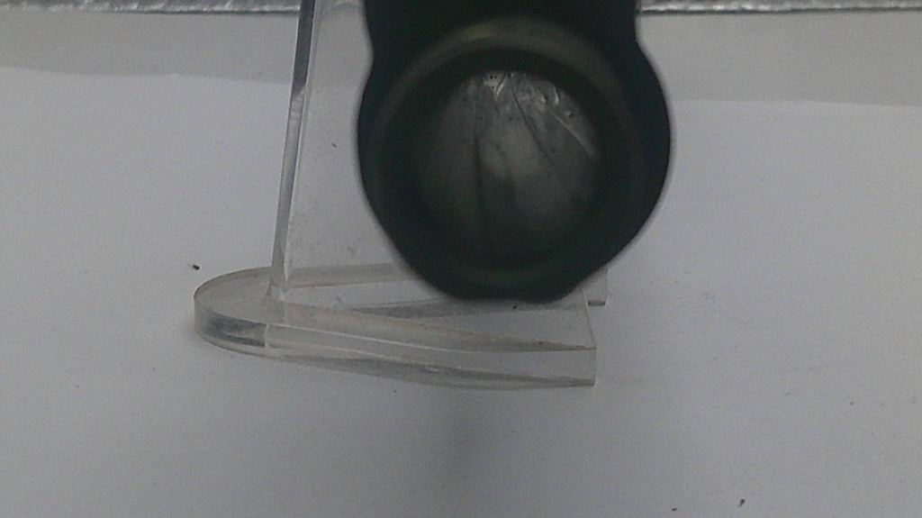A close-up of a black cylindrical object, possibly a lens or filter, positioned in front of a clear acrylic stand on a white surface.