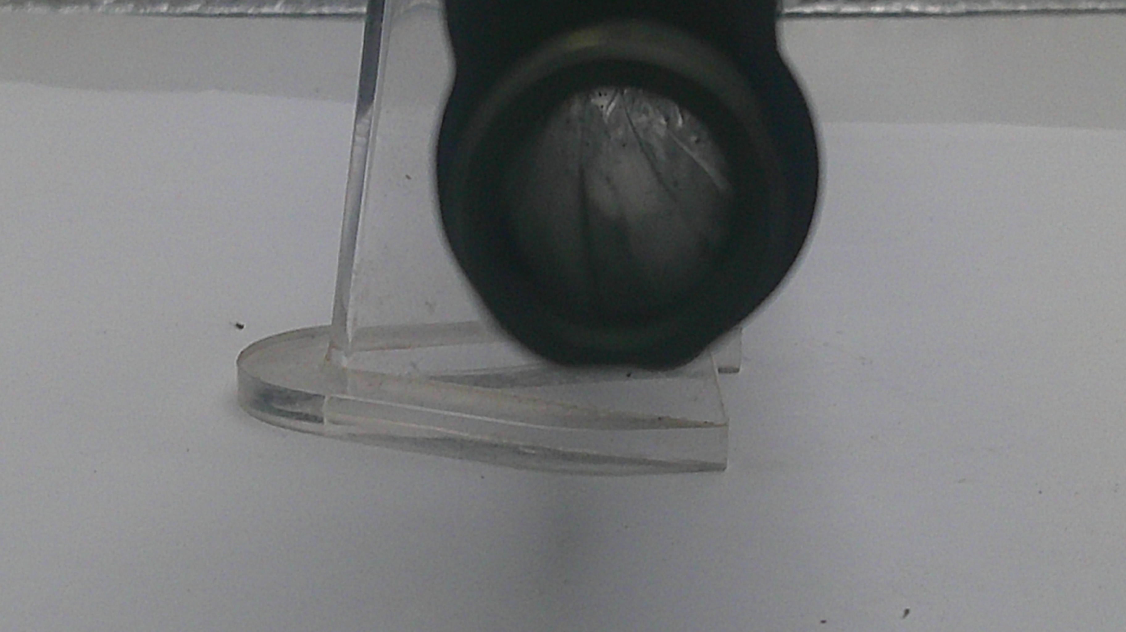 A close-up of a black cylindrical object, possibly a lens or filter, positioned in front of a clear acrylic stand on a white surface.