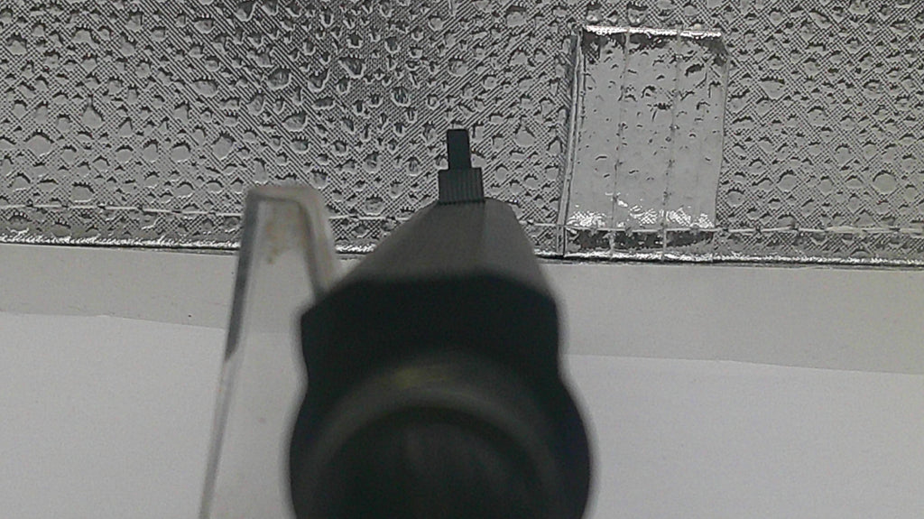 A close-up shot of a black plastic nozzle or cap in the foreground, with a textured silver metallic surface and a white background behind it.