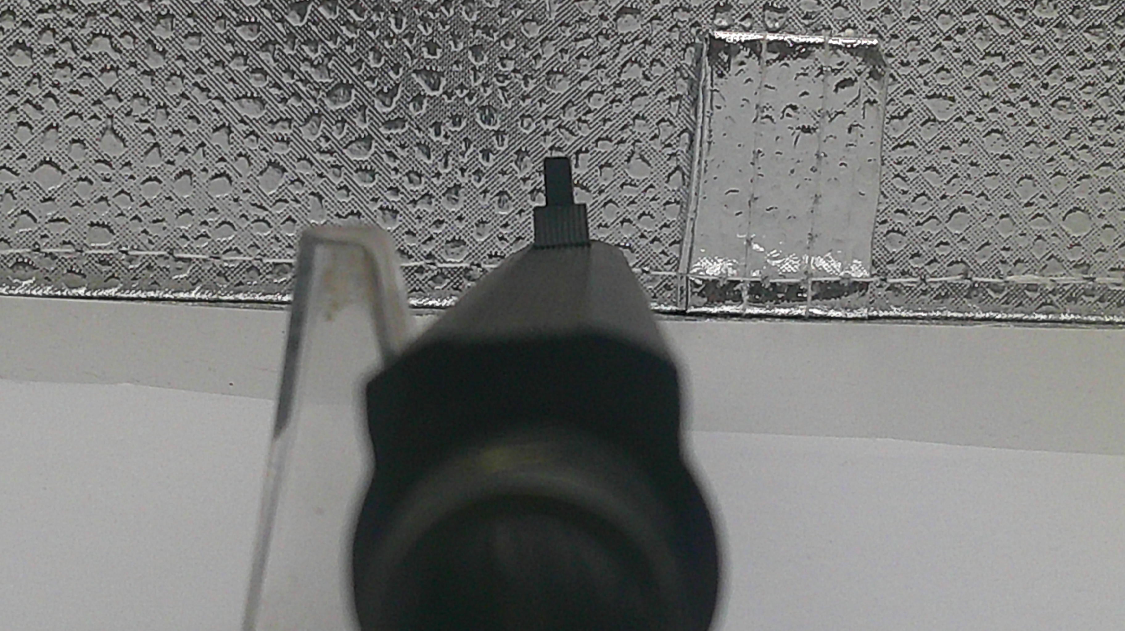 A close-up shot of a black plastic nozzle or cap in the foreground, with a textured silver metallic surface and a white background behind it.
