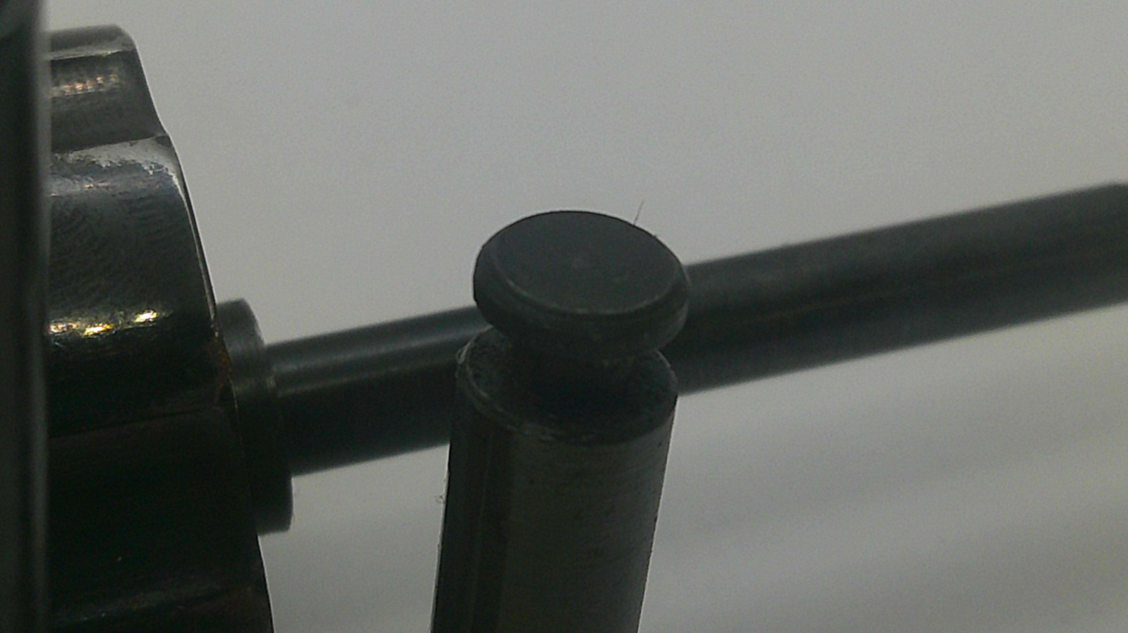 Close-up of a black metal mechanical component with a cylindrical post and horizontal rod against a plain light background.