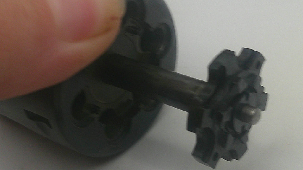A close-up of a small black plastic gear-like component attached to a dark rod, held by a human finger against a plain light background.