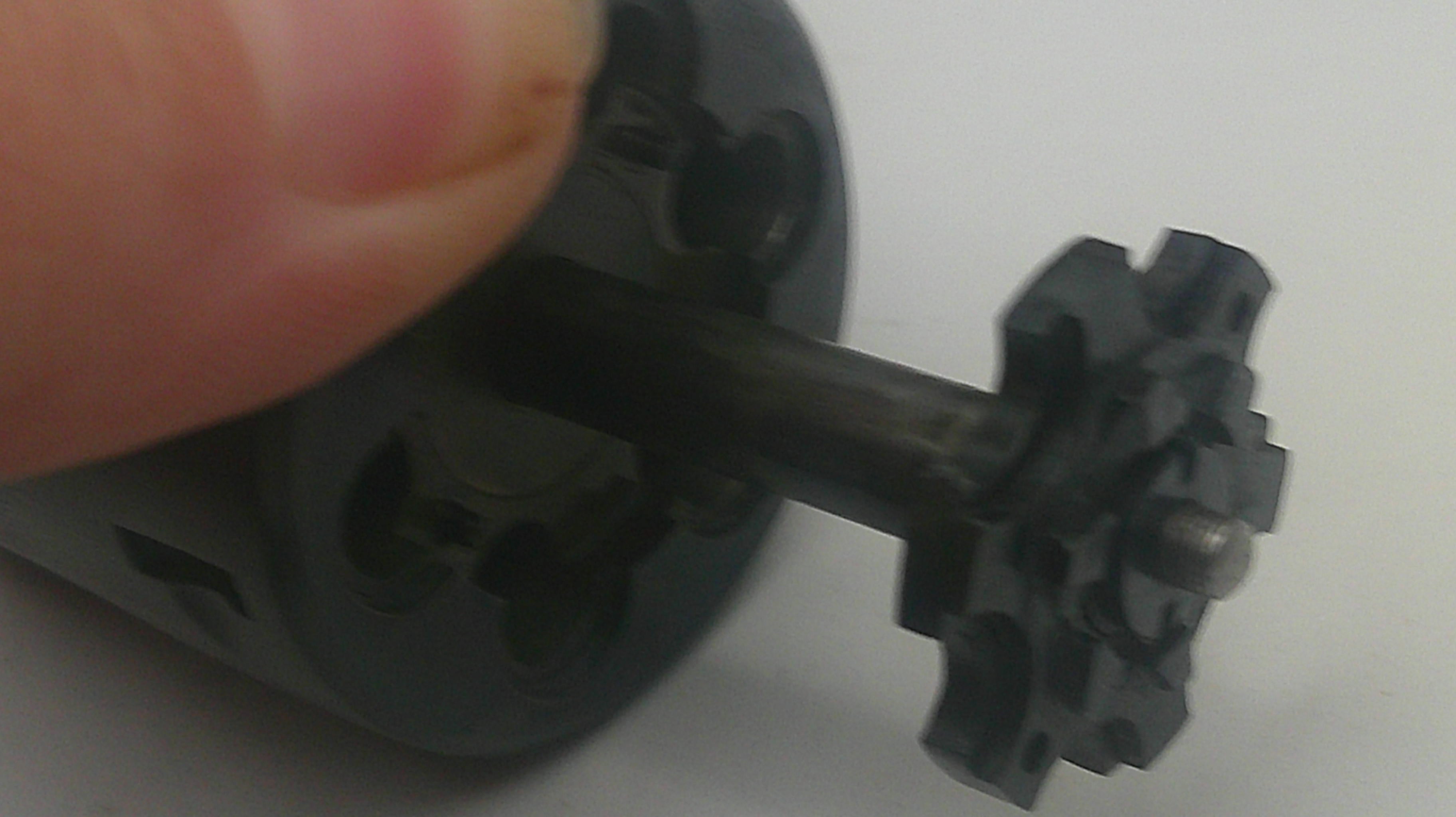A close-up of a small black plastic gear-like component attached to a dark rod, held by a human finger against a plain light background.