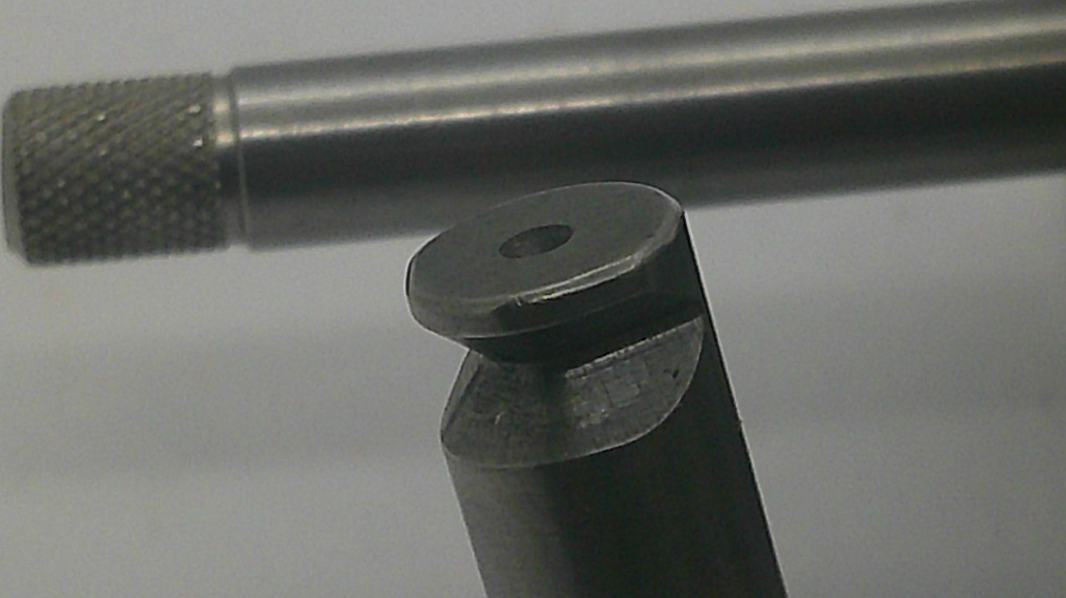 Close-up of a dark metallic cylindrical tool or hardware piece with a knurled textured end, shown against a plain light background.