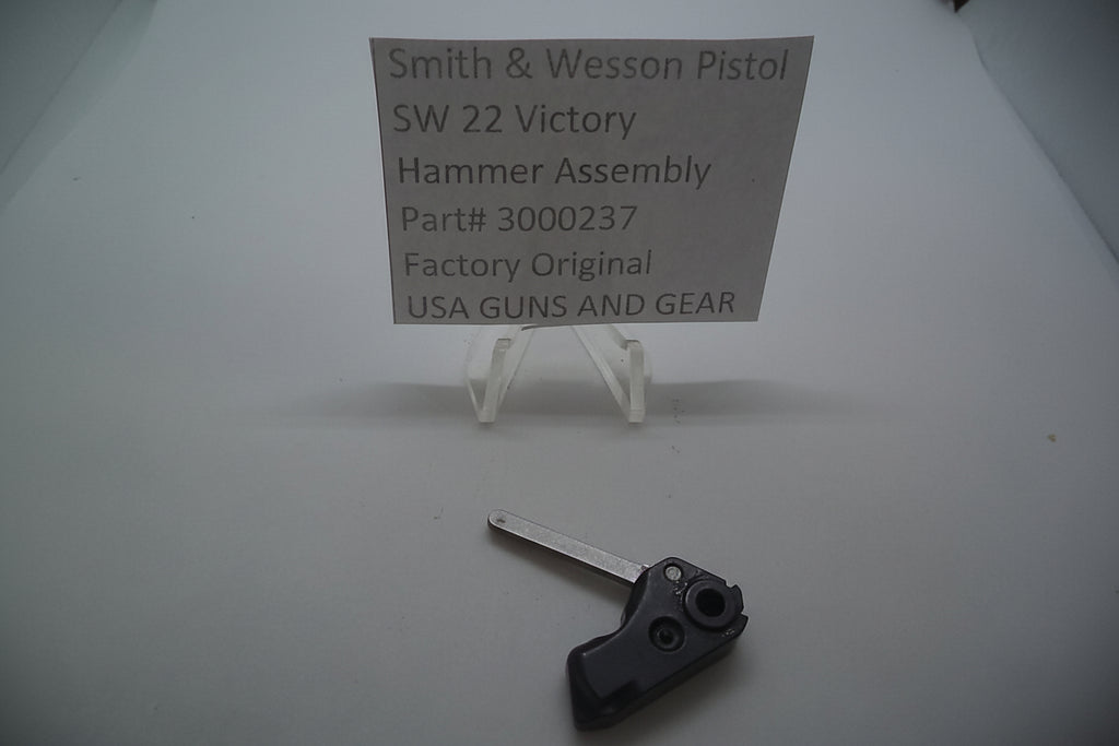 Smith & Wesson SW22 Victory Hammer Assembly displayed on a tripod stand with a sign indicating the part number and manufacturer, accompanied by a key for size comparison.